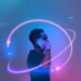 A person holds a smartphone to their ear, standing against a futuristic blue and purple backdrop, reminiscent of Ziimp.com Tech Trends 2025. Neon light trails encircle them, creating a dynamic, techno-inspired atmosphere as digital patterns and data symbols adorn the scene. | ZIIMP