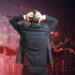 A person in a suit stands with hands on head, facing a large digital screen with declining financial graphs and red trends, symbolizing the challenge of overcoming emotional investing and staying rational in volatile markets. | ZIIMP