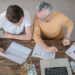 Two people sit at a wooden table with financial charts, a laptop, cash, coins in a jar, and documents, discussing How to Set Smart Financial Goals: A Beginner's Guide to Investing. They are viewed from above, focused on the papers in front of them. | ZIIMP