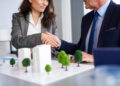 Two businesspeople in suits shake hands over a desk with an architectural model of buildings and trees, suggesting a real estate or business agreement. | ZIIMP