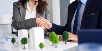 Two businesspeople in suits shake hands over a desk with an architectural model of buildings and trees, suggesting a real estate or business agreement. | ZIIMP