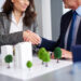 Two businesspeople in suits shake hands over a desk with an architectural model of buildings and trees, suggesting a real estate or business agreement. | ZIIMP
